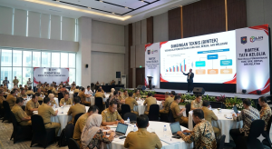 Suasana acara Bimbingan Teknis (Bimtek) Tata Kelola Pemerintahan di dalam aula besar. Puluhan peserta berseragam dinas duduk mengitari meja-meja bundar sambil memperhatikan presentasi narasumber di panggung. Layar LED besar di depan menampilkan materi tentang tata kelola pemerintahan yang baik, bersih, dan melayani.