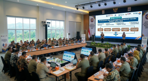 Foto suasana Bimtek/Pelatihan Perencanaan dan Penganggaran Responsif Gender (PPRG) di sebuah aula pertemuan. Puluhan peserta berseragam dinas PNS dan batik duduk dalam formasi meja berbentuk U, fokus memperhatikan narasumber di depan. Di atas panggung, seorang narasumber wanita sedang memberikan paparan di depan layar LED besar yang menampilkan materi "Perencanaan dan Penganggaran Responsif Gender (PPRG) Tahun 2026", lengkap dengan bagan alur proses, logo KPPPA, Bappenas, dan LKPP. Ruangan terlihat modern dengan pencahayaan yang terang dan pemandangan luar melalui jendela kaca besar.