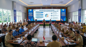 Foto suasana Bimtek/Pelatihan Dasar: Kedisiplinan dan Kapasitas Aparatur di dalam aula. Para peserta berseragam dinas PNS (khaki) dan batik duduk rapi dalam formasi meja berbentuk U. Di depan ruangan, dua orang narasumber (pria dan wanita) berdiri memandu acara dengan latar belakang layar LED besar berlatar putih-biru yang menampilkan poin materi seperti Nilai-Nilai ASN, Etika Birokrasi, dan Disiplin Kerja.