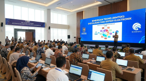 Suasana acara Bimbingan Teknis (Bimtek) Digitalisasi dan Sistem Elektronik yang dihadiri oleh puluhan peserta di dalam aula besar. Di depan ruangan, terdapat layar LED besar yang menampilkan materi "Sistem Pemerintahan Berbasis Elektronik (SPBE)" dengan logo Kementerian Dalam Negeri dan Kominfo, serta seorang narasumber yang sedang memberikan presentasi di panggung. Para peserta tampak duduk rapi menghadap panggung sambil membuka laptop masing-masing.