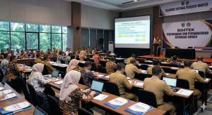 Foto suasana acara Bimbingan Teknis (Bimtek) Perencanaan dan Penganggaran Keuangan Daerah yang dihadiri oleh ratusan peserta pemerintah daerah di dalam sebuah aula besar. Di depan ruangan, seorang narasumber yang mengenakan kemeja batik sedang memberikan presentasi dari atas panggung menggunakan proyektor besar. Layar presentasi menampilkan materi bertuliskan 'BIMBINGAN TEKNIS (BIMTEK)' dan 'PERENCANAAN DAN PENGANGGARAN KEUANGAN DAERAH' dengan diagram alur di bawahnya. Peserta yang terdiri dari pria dan wanita terlihat duduk rapi di meja-meja panjang, membuka laptop dan buku catatan sambil menyimak materi dengan fokus. Spanduk bertuliskan 'BIMTEK PERENCANAAN DAN PENGANGGARAN' juga terpasang di samping panggung."