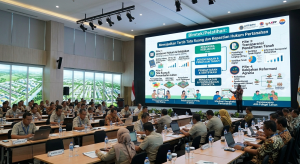 Foto suasana Bimtek/Pelatihan Kebijakan dan Regulasi Agraria di dalam sebuah aula modern. Puluhan peserta berseragam dinas PNS (khaki) dan batik duduk rapi menghadap meja panjang yang dilengkapi laptop. Di panggung depan, seorang narasumber pria berdiri menjelaskan materi yang ditampilkan pada layar LED raksasa. Layar tersebut memuat infografis bertuliskan "Mewujudkan Tertib Tata Ruang dan Kepastian Hukum Pertanahan" serta logo ATR/BPN, BNPB, dan LKPP.